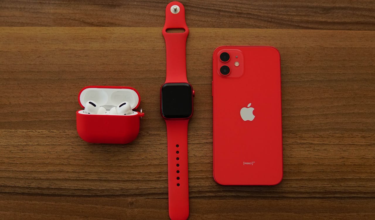 about-us Red Apple iPhone, AirPods, and Apple Watch arranged on a wooden table.